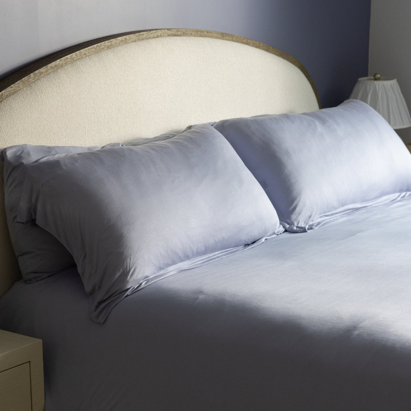 Bedroom with a bed featuring light blue bedding and pillows, a nightstand with a lamp, and a lamp on the wall.