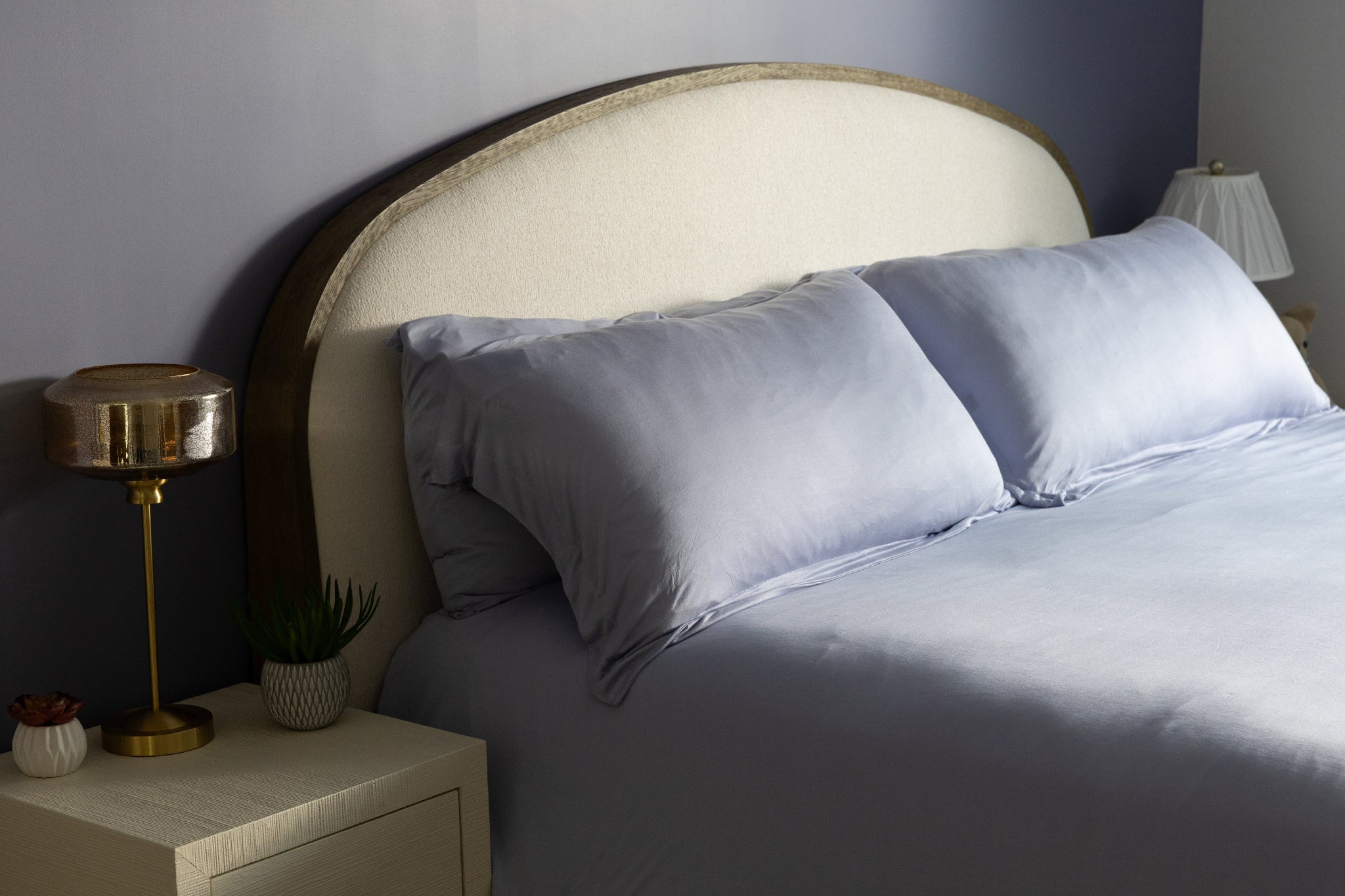 Bedroom with a bed featuring light blue bedding and pillows, a nightstand with a lamp, and a lamp on the wall.