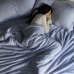 Person lying in bed under a blue comforter with pillows from top down