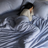 Person lying in bed under a blue comforter with pillows from top down