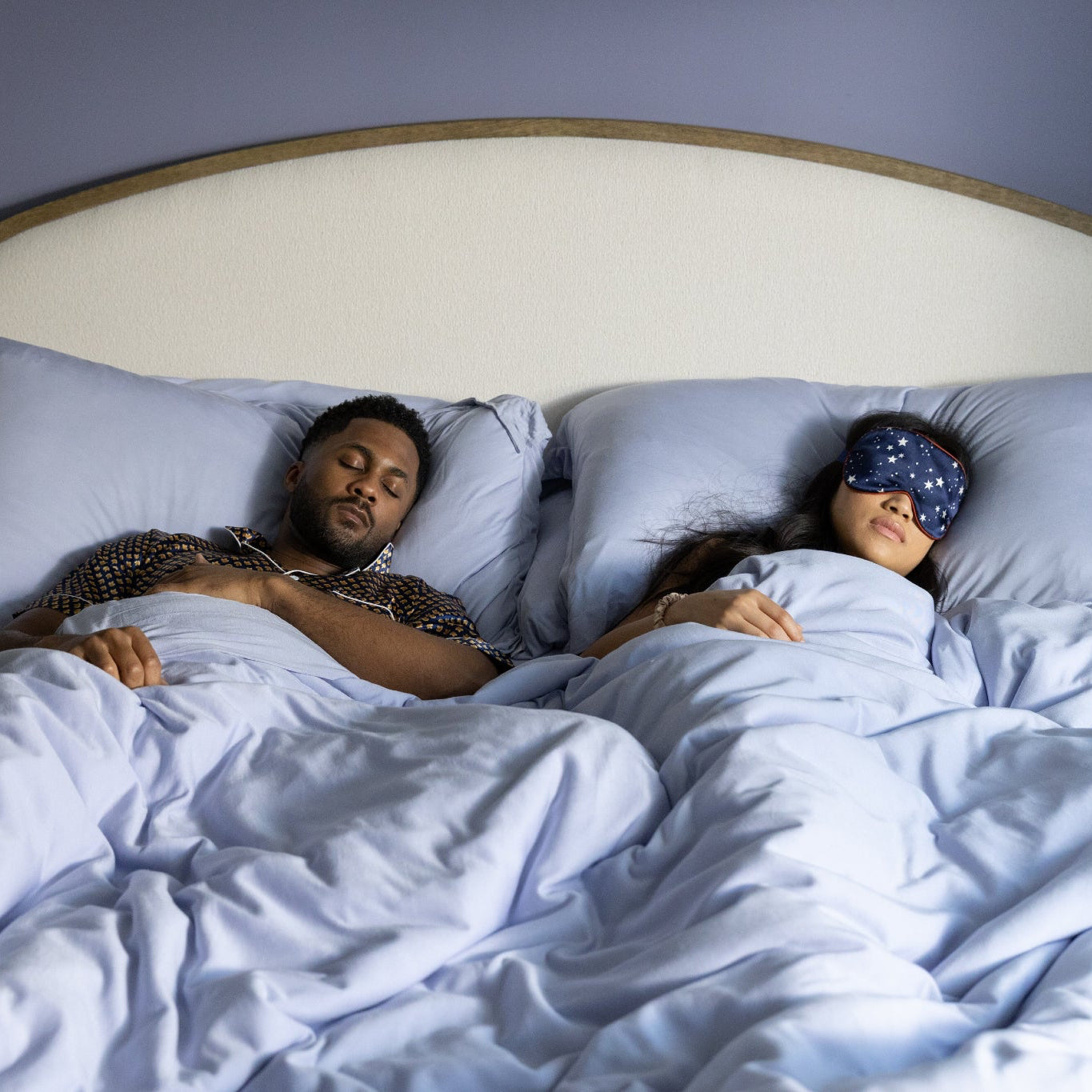 Two people lying in bed, one awake and the other with a sleep mask on.
