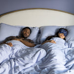 Two people lying in bed, one awake and the other with a sleep mask on.
