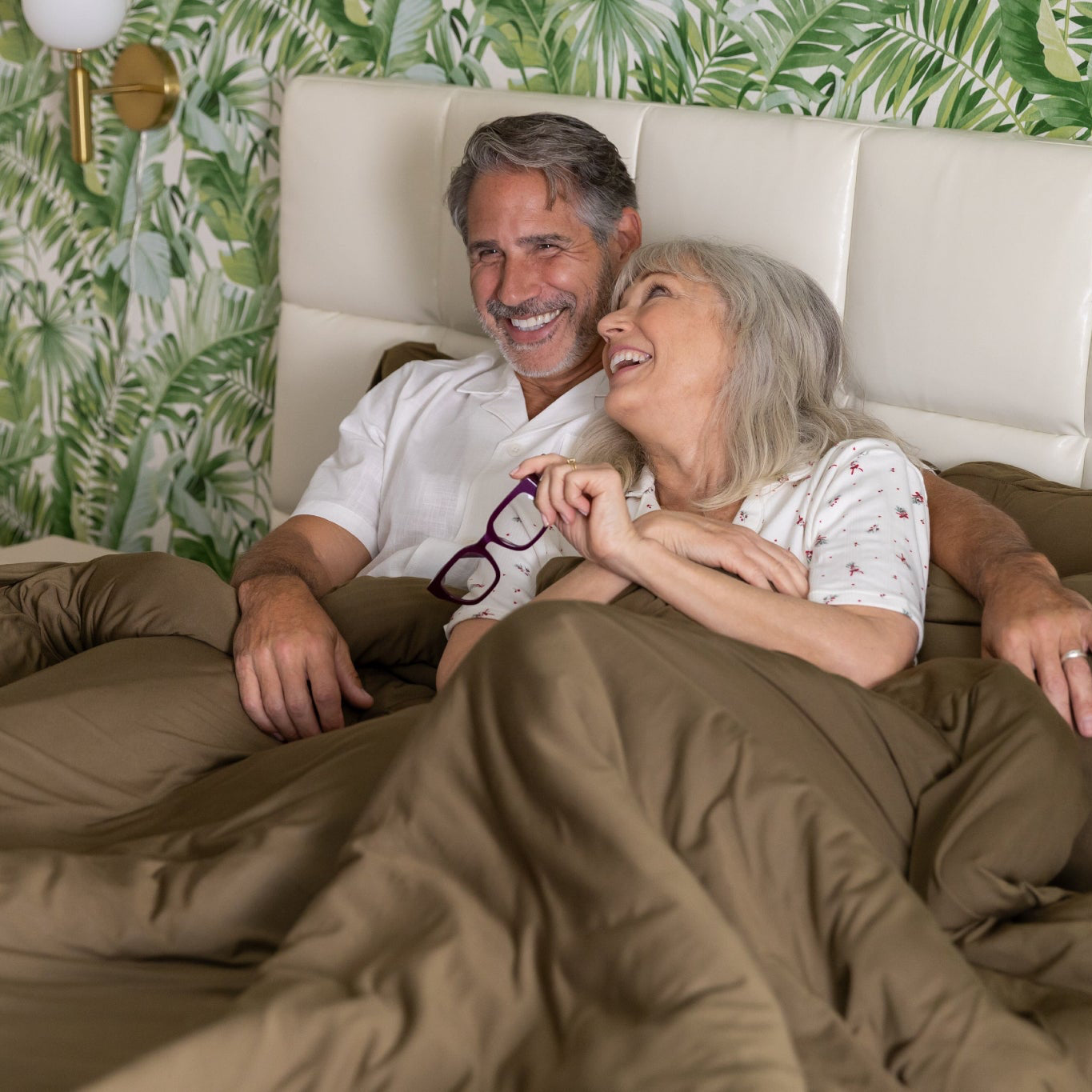older Couple lying in bed together with a tropical-themed wallpaper.