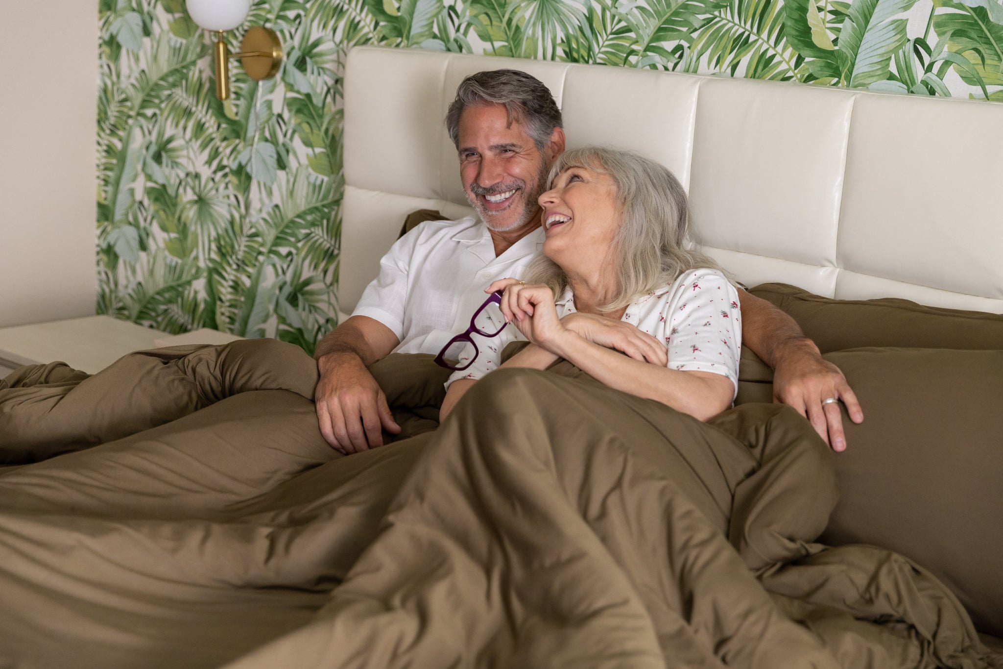older Couple lying in bed together with a tropical-themed wallpaper.