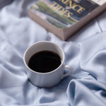 White mug with coffee on a soft blue fabric surface, with a book titled 'Prince Tides' partially visible.