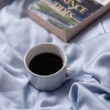 White mug with coffee on a soft blue fabric surface, with a book titled 'Prince Tides' partially visible.