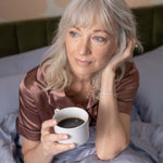 Woman in bed holding a cup of coffee with a neutral background