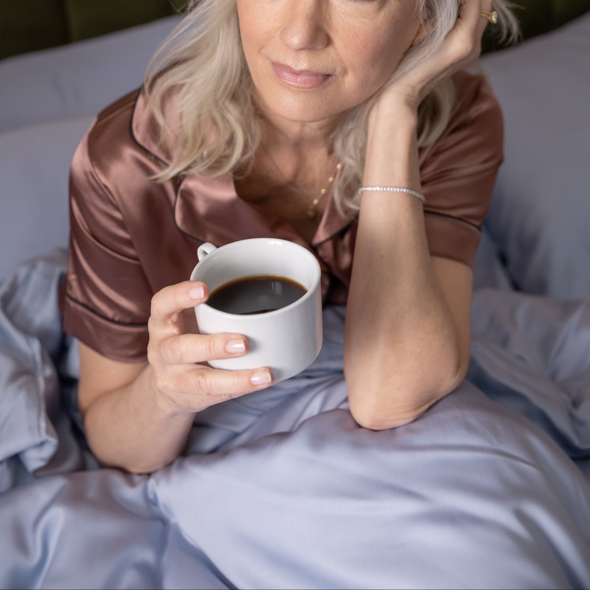 Woman in bed holding a cup of coffee with a neutral background