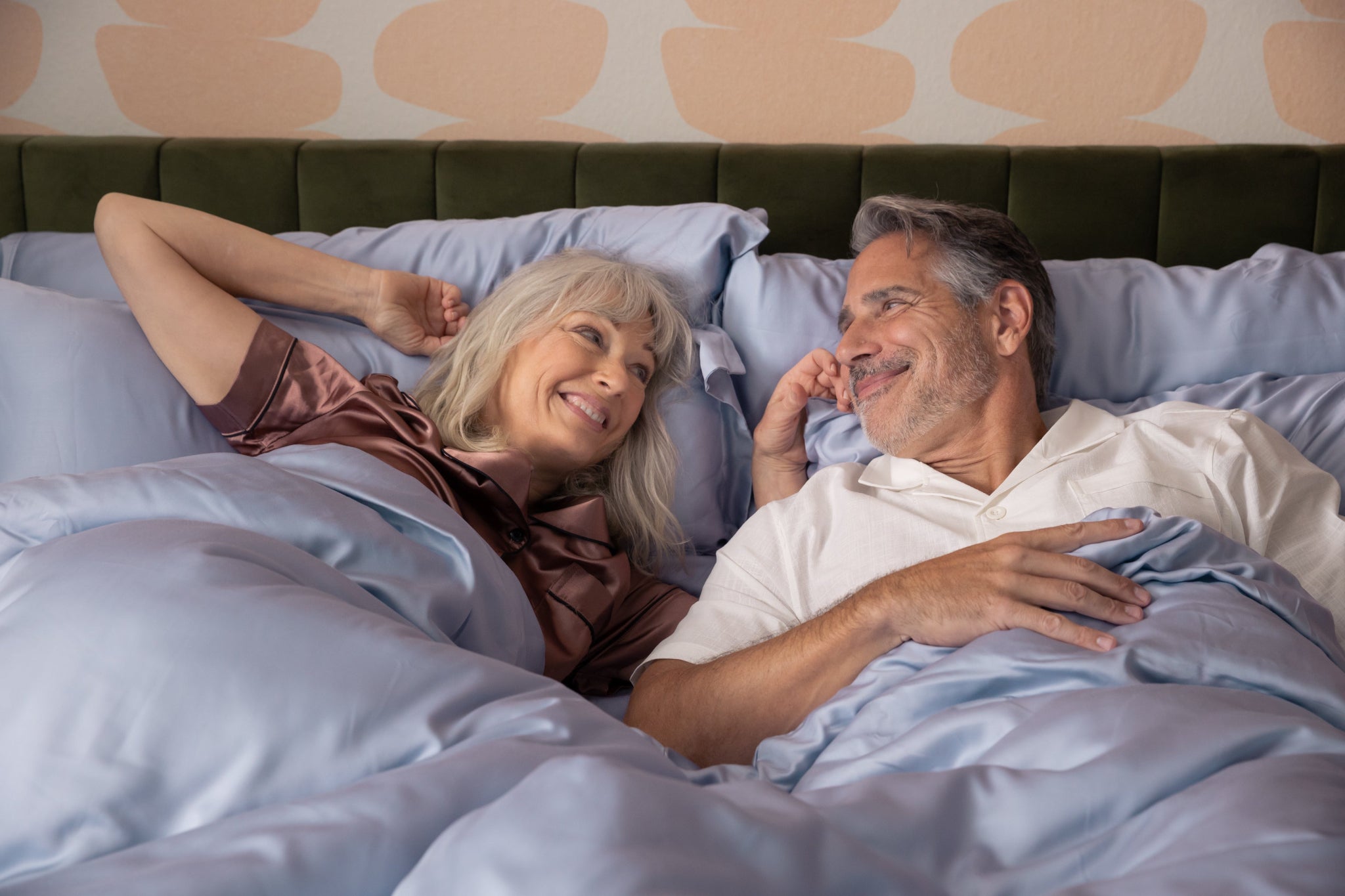 Couple lying in bed together, smiling and embracing on dream blue sateen sheets